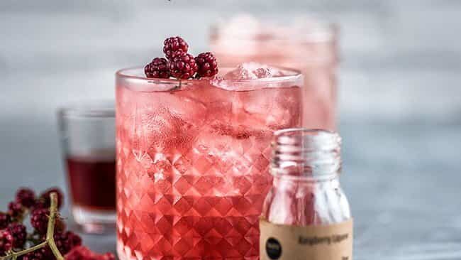 A glass of pink berry cocktail with ice, garnished with fresh berries. A small bottle and another drink are in the background, along with loose berries on the table. The scene is bright and refreshing.