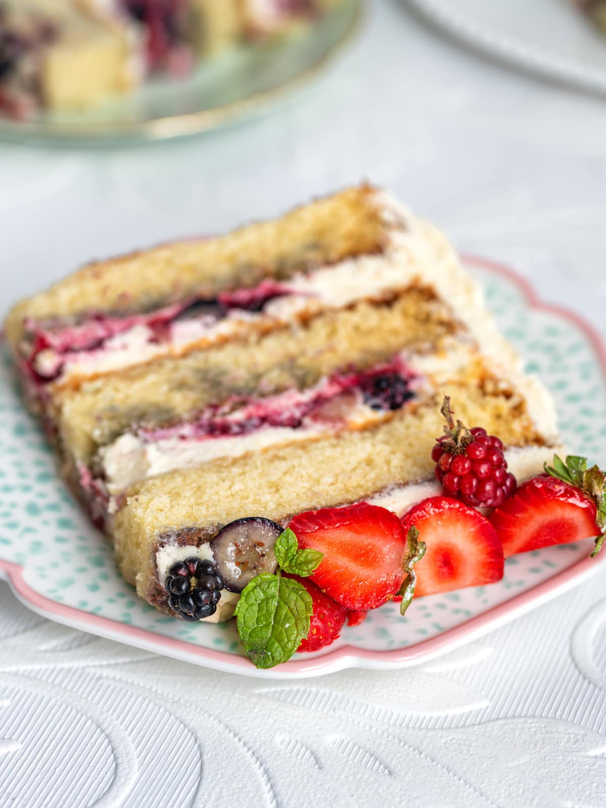 A slice of Berry Chantilly cake with cream and berries is on a plate, garnished with fresh strawberries, mint leaves, and blackberries.