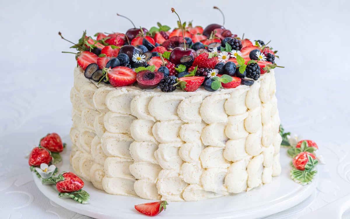 A tall chantilly cake with fresh berries features textured white frosting, crowned with a medley of strawberries, cherries, blueberries, blackberries, and delicate small flowers. It sits on a white plate adorned with sliced strawberries and greenery around the base.