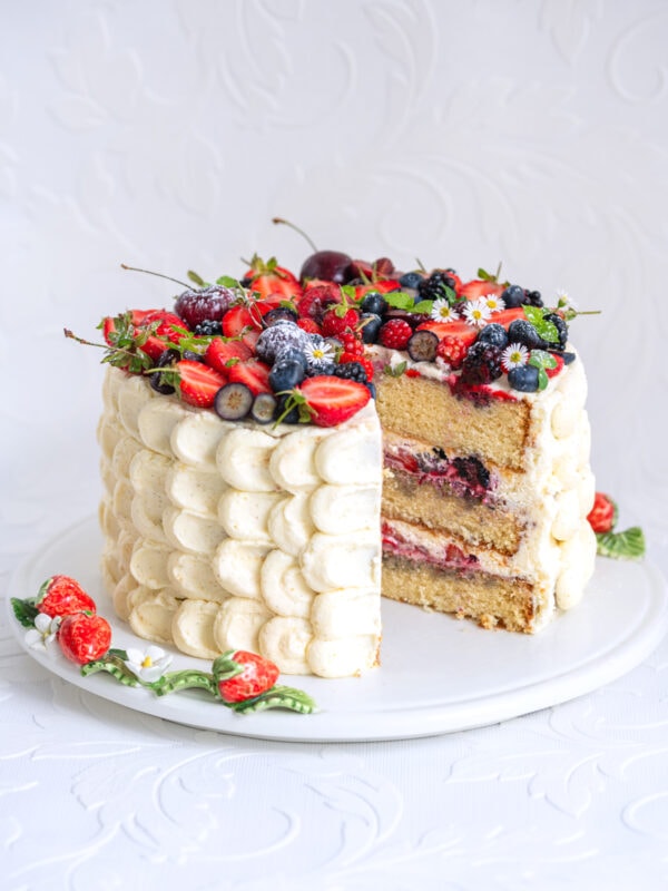 A tall, layered vanilla cake with scalloped white frosting, topped with assorted berries and small flowers. A large slice has been removed, showing fruit filling between the layers. The cake sits on a white plate.