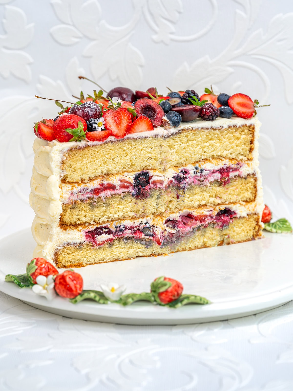 A three-layer Berry Chantilly Cake with cream and mixed berries between layers is topped with fresh strawberries, cherries, blueberries, and small white flowers, all elegantly arranged on a white plate against a white background.