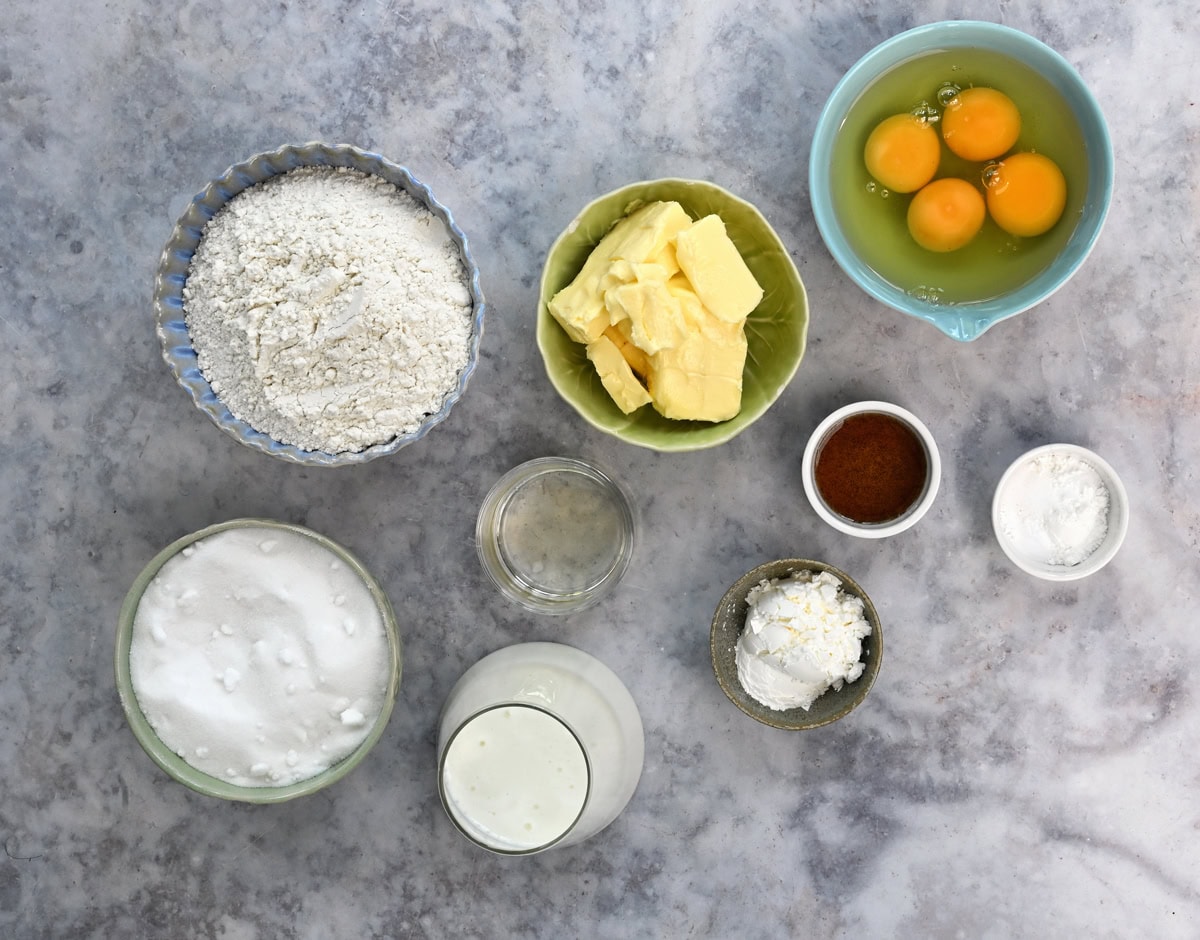 Baking ingredients on a gray marble surface: eggs, flour, butter, sugar, milk, cornstarch, baking powder, vanilla, and a small empty glass jar, each in separate bowls or cups, arranged neatly from above.