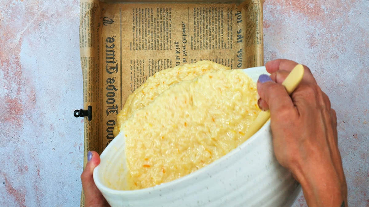 A person pours a mixture from a white bowl into a rectangular baking pan lined with parchment paper printed with newspaper-like text.