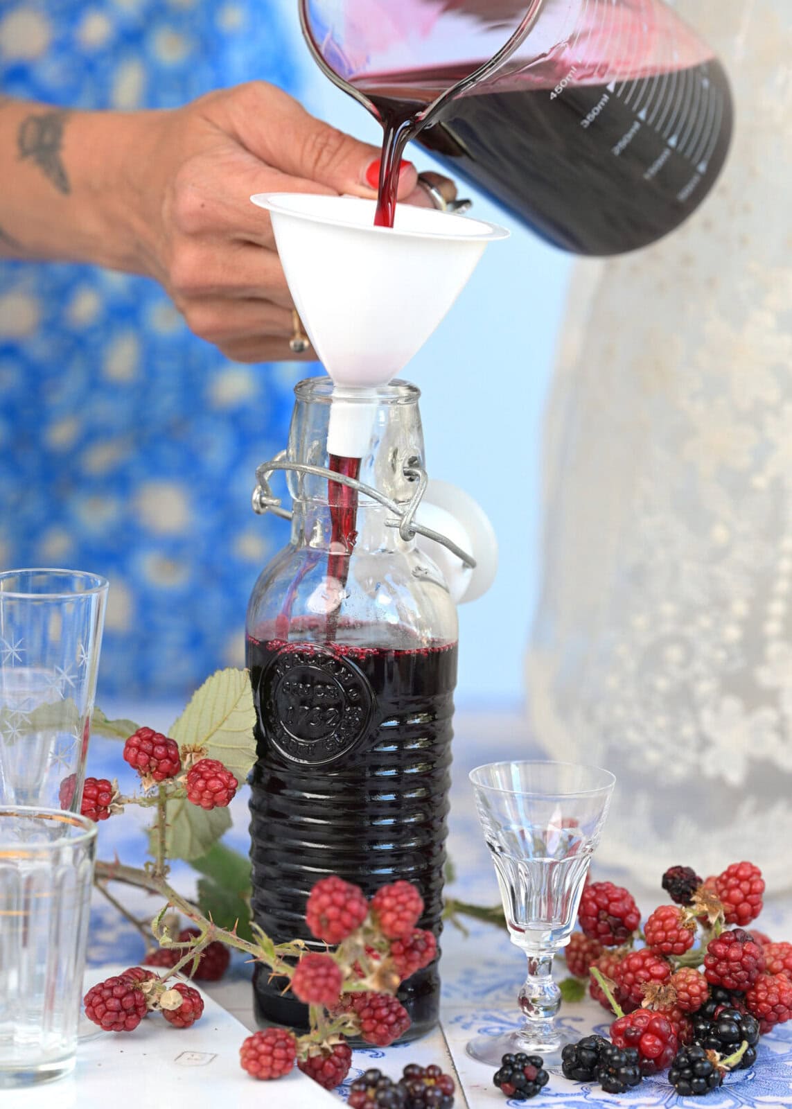 A person pours dark berry juice through a funnel into a glass bottle. Fresh blackberries and glasses are on the table. Another person stands nearby, both wearing light-colored clothing.