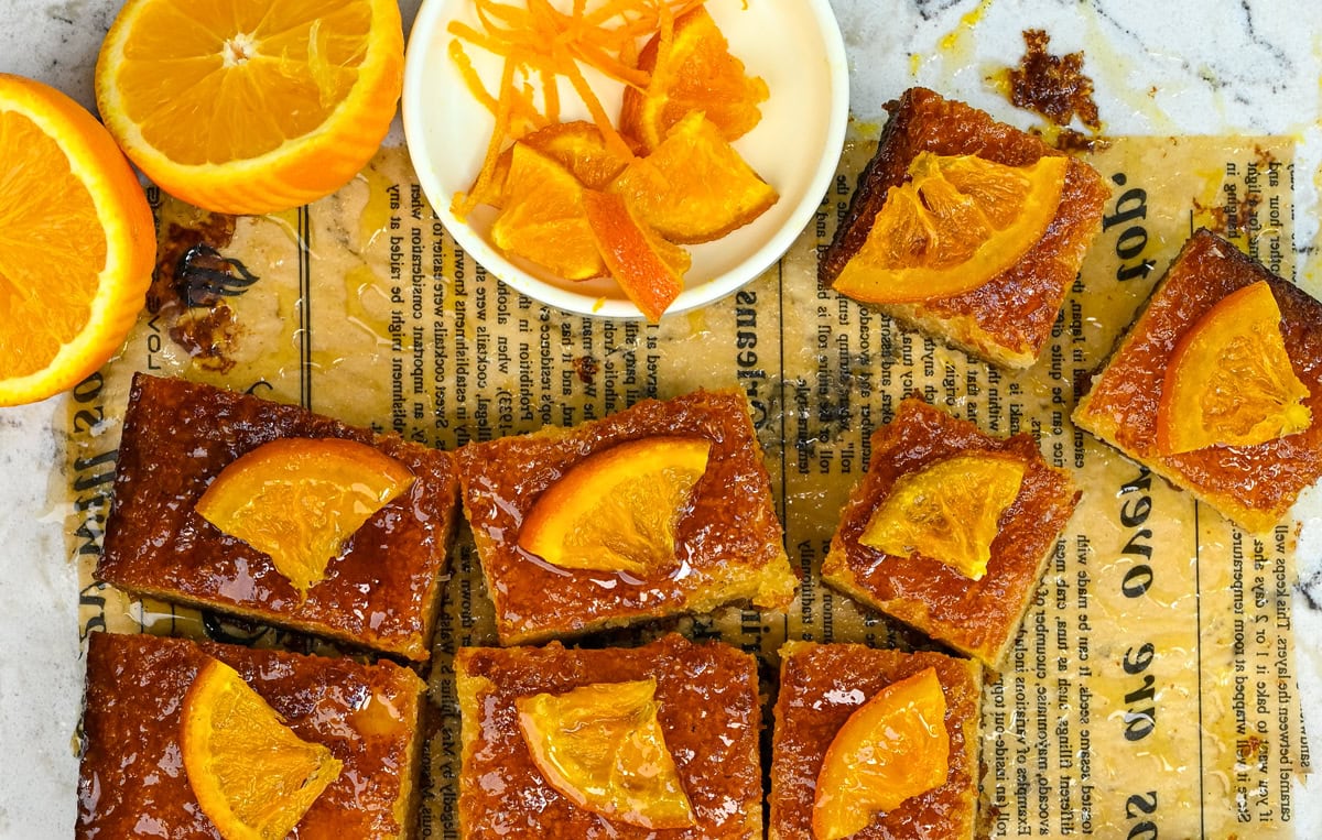 Nine pieces of Greek orange cake topped with orange slices, arranged on parchment paper with text, next to a sliced orange and a small dish holding more orange slices and zest.