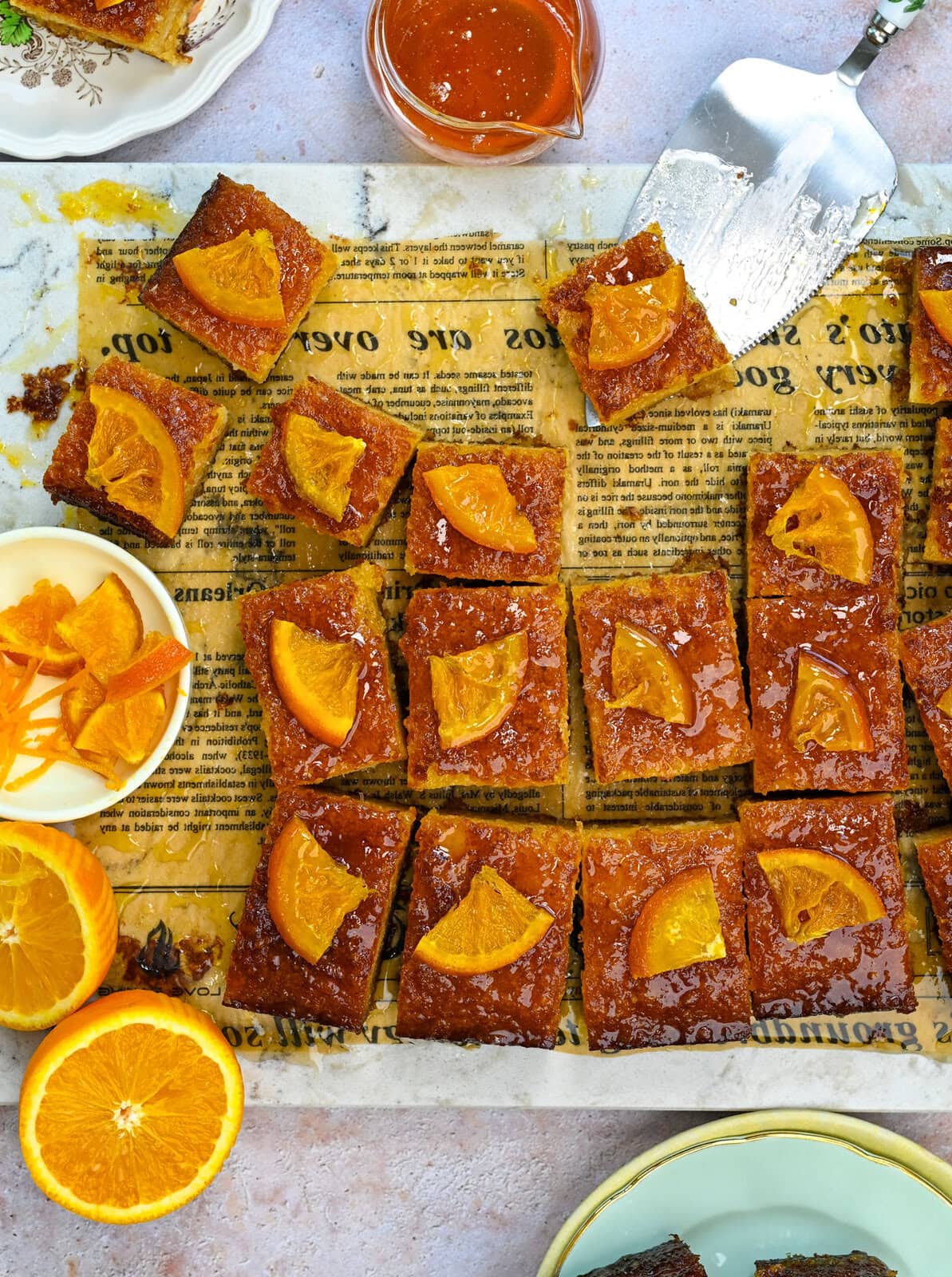 Orange-glazed portokalopita cake squares topped with orange slices are arranged on a sheet of printed parchment. Fresh orange slices, zest, and a bowl of glaze are placed nearby for decoration, highlighting the vibrant flavors of this Greek orange phyllo cake.