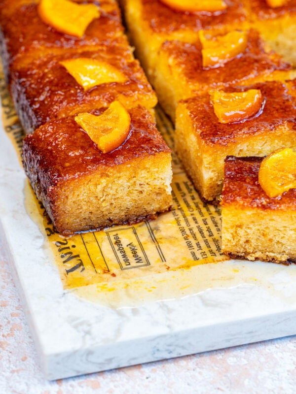 Golden, square slices of moist portokalopita orange phyllo cake topped with glossy peach pieces are arranged on a marble slab with parchment paper. One slice is slightly pulled out, showing the cake’s fluffy texture and syrupy glaze.