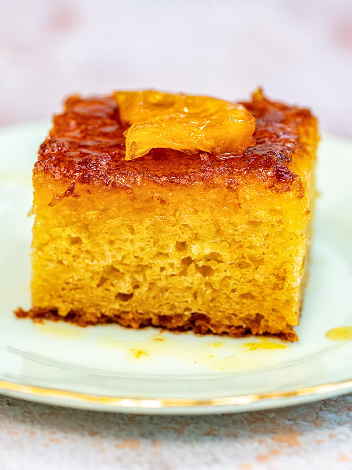 A close-up of a square piece of portokalopita (orange phyllo cake) with a candied orange slice on top, served on a white plate with a gold rim. The cake appears moist and golden brown.