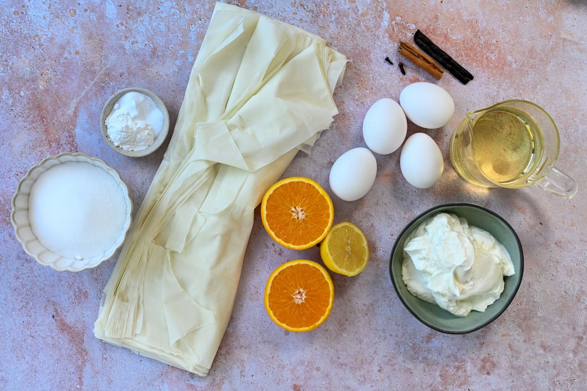 Various baking ingredients are arranged on a countertop, including phyllo dough, eggs, sugar, yogurt, orange and lemon halves, cinnamon sticks, vanilla beans, cornstarch, and a cup of oil.