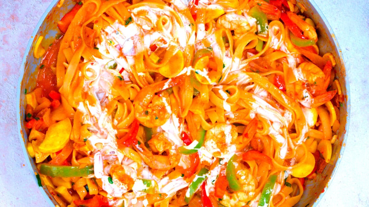 Colorful pasta with shrimp, sliced bell peppers, and a creamy sauce mixed together in a large pan. The dish is vibrant with orange, red, green, and yellow ingredients visible throughout.