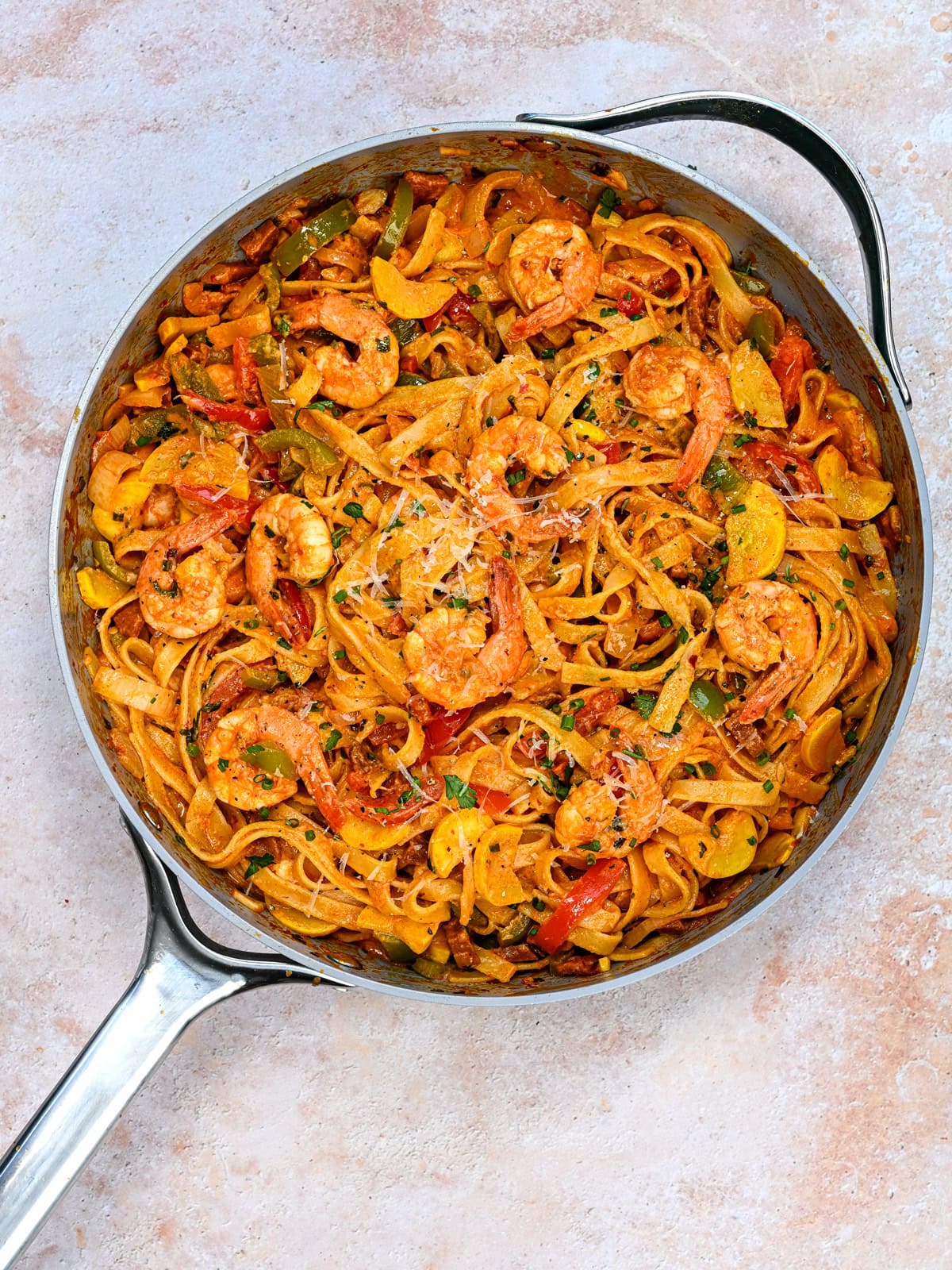 A skillet filled with creamy shrimp fettuccine with chorizo, bell peppers, and herbs, topped with grated cheese, sits on a light-colored surface.