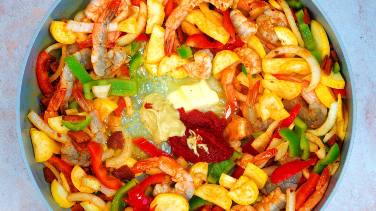 Colorful pan filled with raw shrimp, sliced bell peppers, onions, squash, chorizo, butter, mustard, and tomato paste, ready to be cooked.