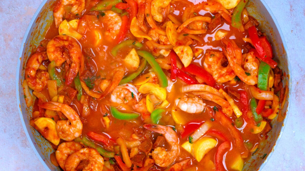 Shrimp, bell peppers, onions, and squash are simmered in a rich orange-red sauce, creating a colorful and hearty seafood stew in a pan.