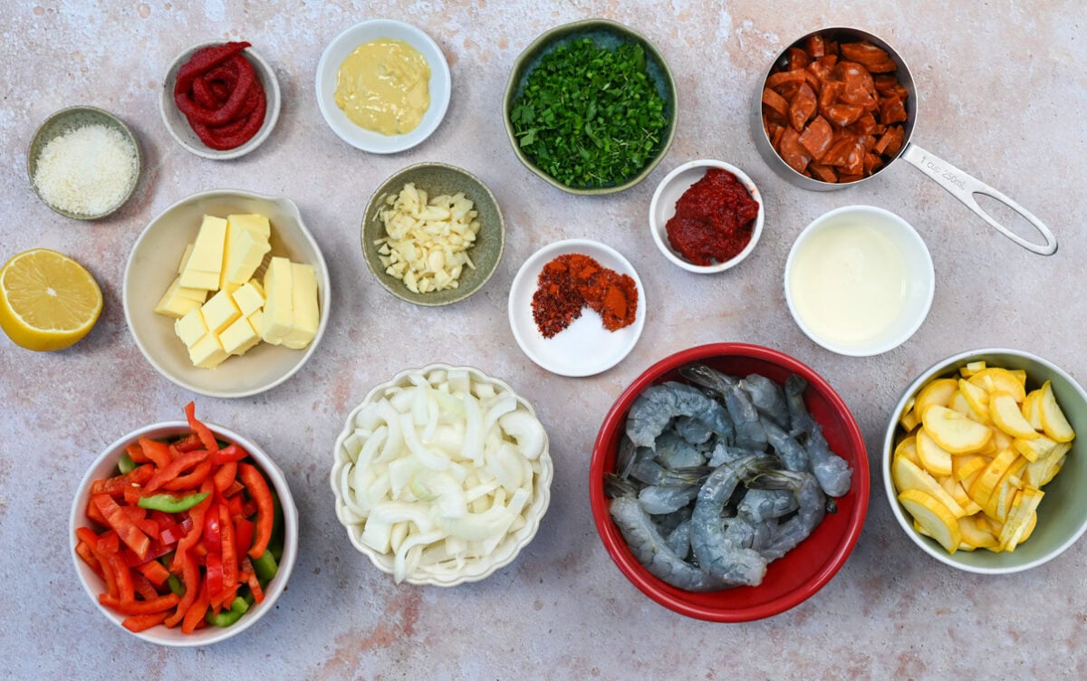 Various ingredients in bowls and cups on a countertop, including chopped onions, bell peppers, shrimp, sliced sausage, lemon, butter, garlic, parsley, spices, cheese, squash, tomato paste, oil, and mustard.