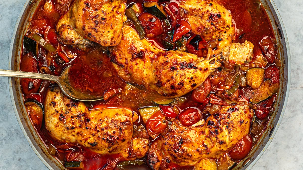 Four browned chicken breasts simmer in a skillet with cherry tomatoes, zucchini, and a rich red sauce. A spoon rests in the pan, and the dish looks freshly cooked and flavorful.