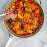 A skillet filled with cooked chicken breasts, cherry tomatoes, zucchini, and herbs in a savory tomato sauce resembles a delightful chicken casserole, with a metal spoon on the side.