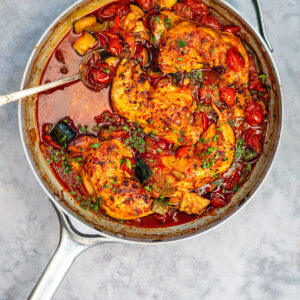 A skillet filled with cooked chicken breasts, cherry tomatoes, zucchini, and herbs in a savory tomato sauce resembles a delightful chicken casserole, with a metal spoon on the side.