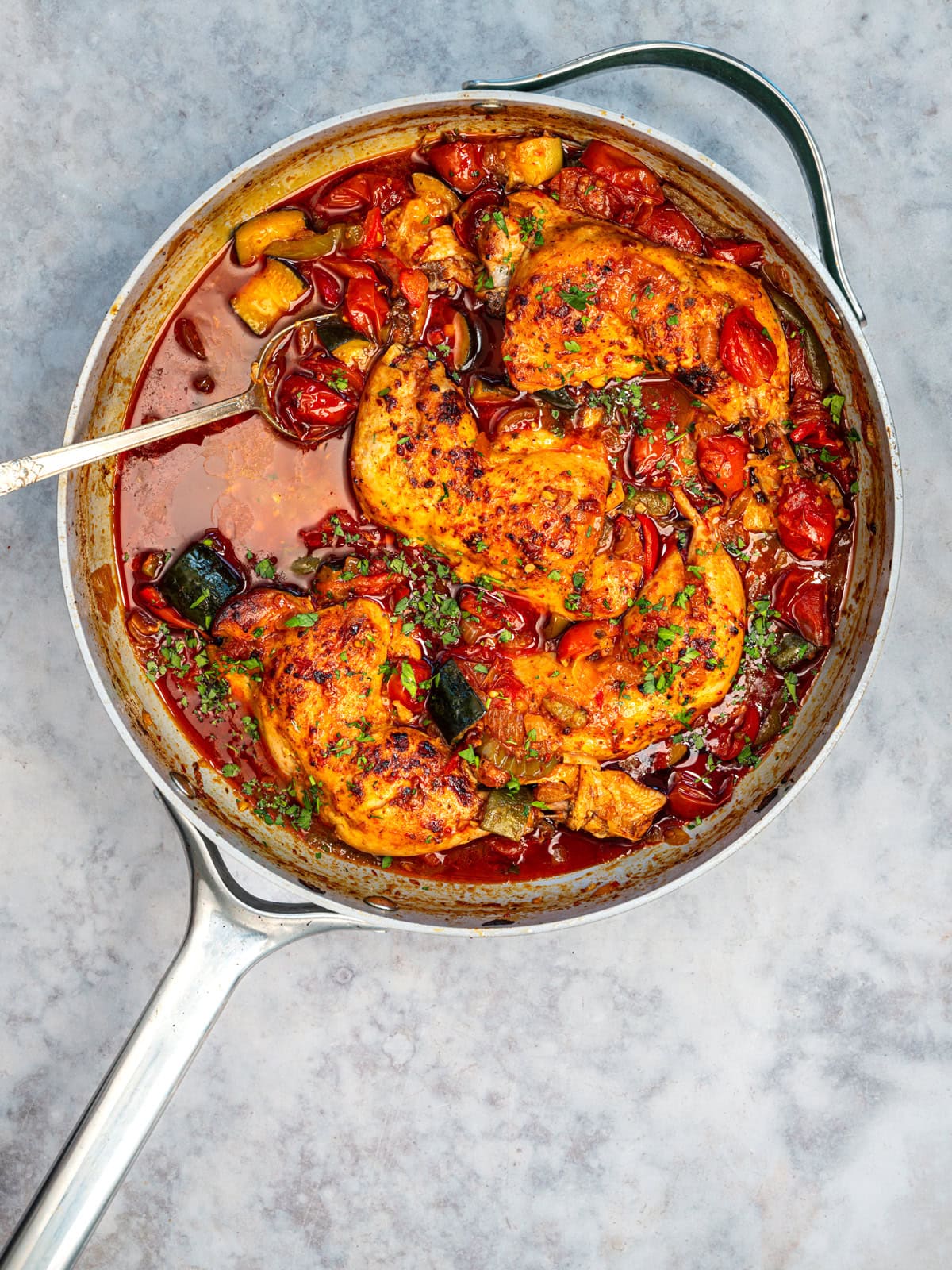 A skillet filled with cooked chicken breasts, cherry tomatoes, zucchini, and herbs in a savory tomato sauce resembles a delightful chicken casserole, with a metal spoon on the side.