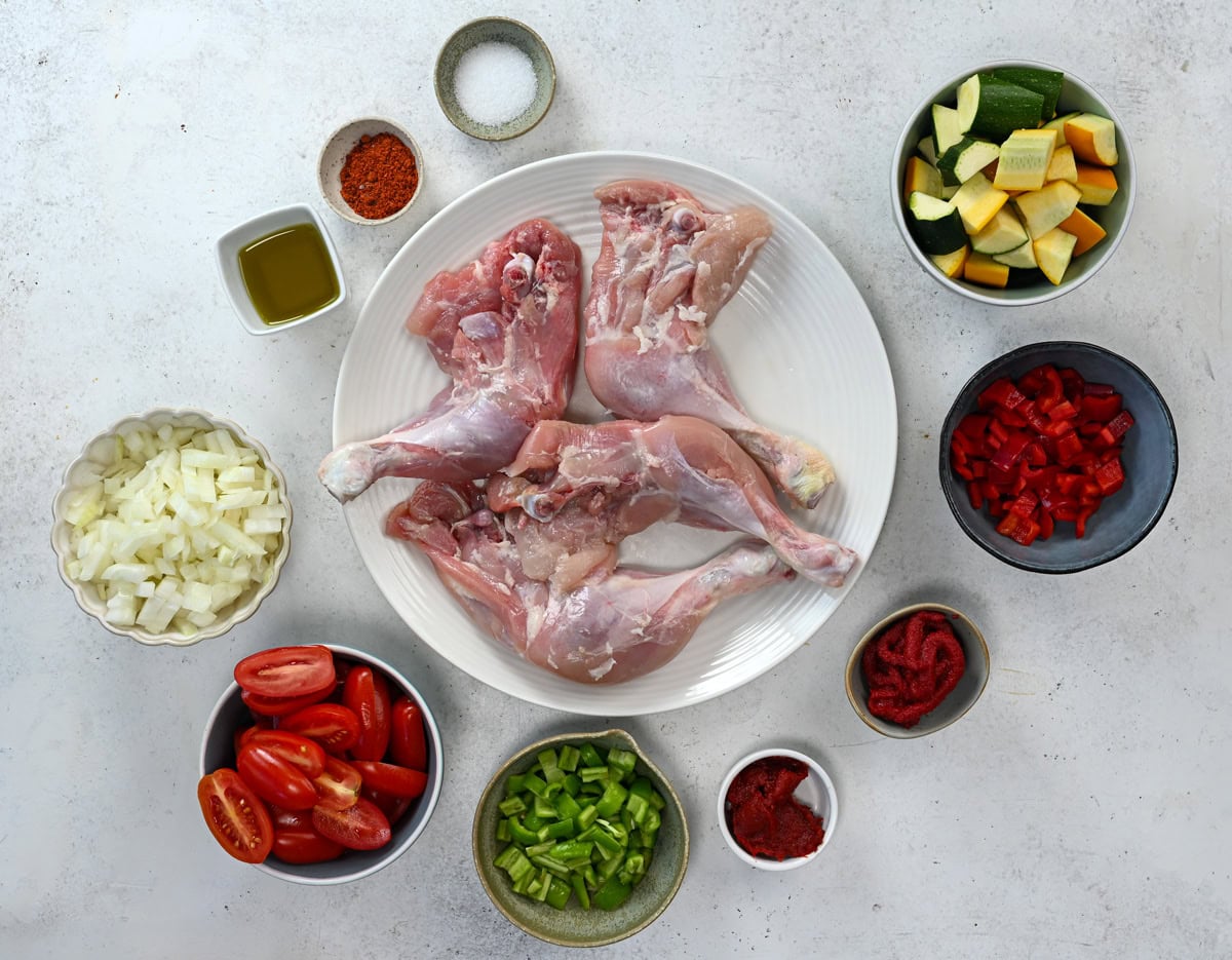 Raw chicken legs on a white plate, surrounded by bowls of chopped onions, bell peppers, tomatoes, zucchini, tomato paste, spices, and a small dish of olive oil, all arranged on a white surface.