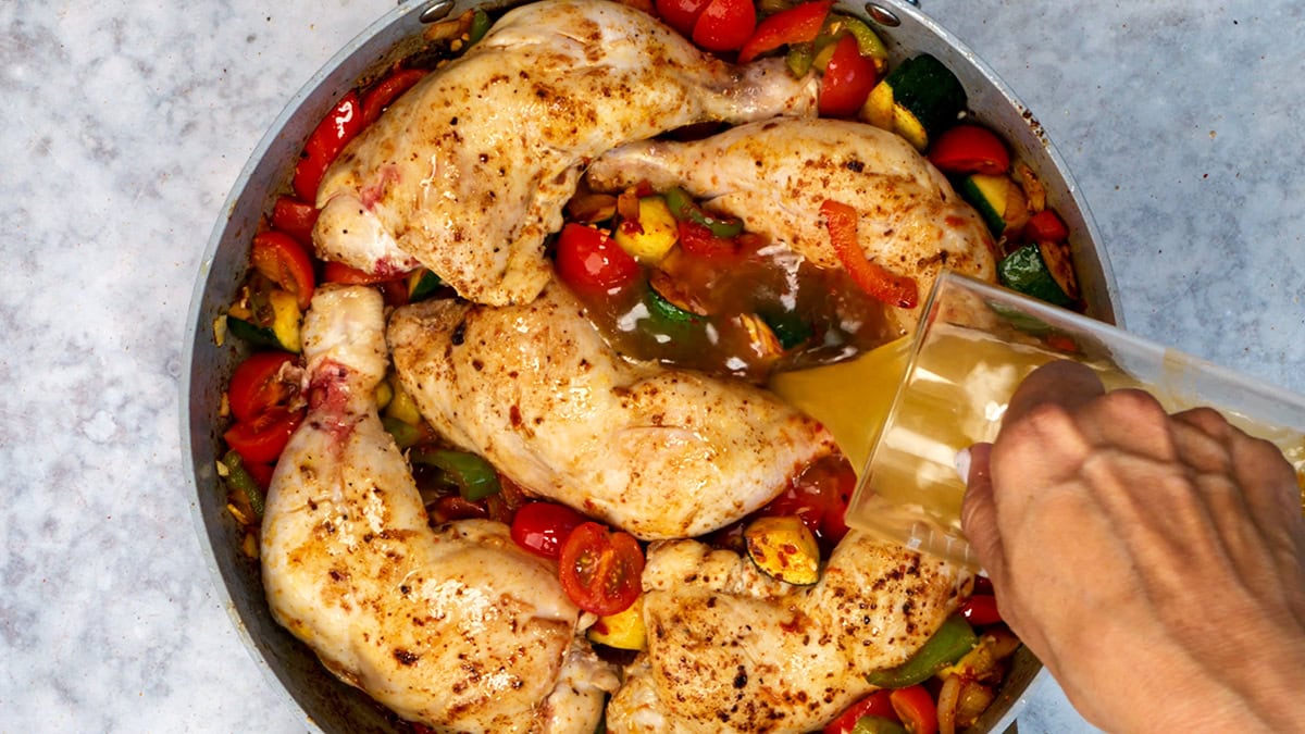 A hand pours broth over seasoned chicken legs in a pan with chopped tomatoes, zucchini, and bell peppers on a light countertop.