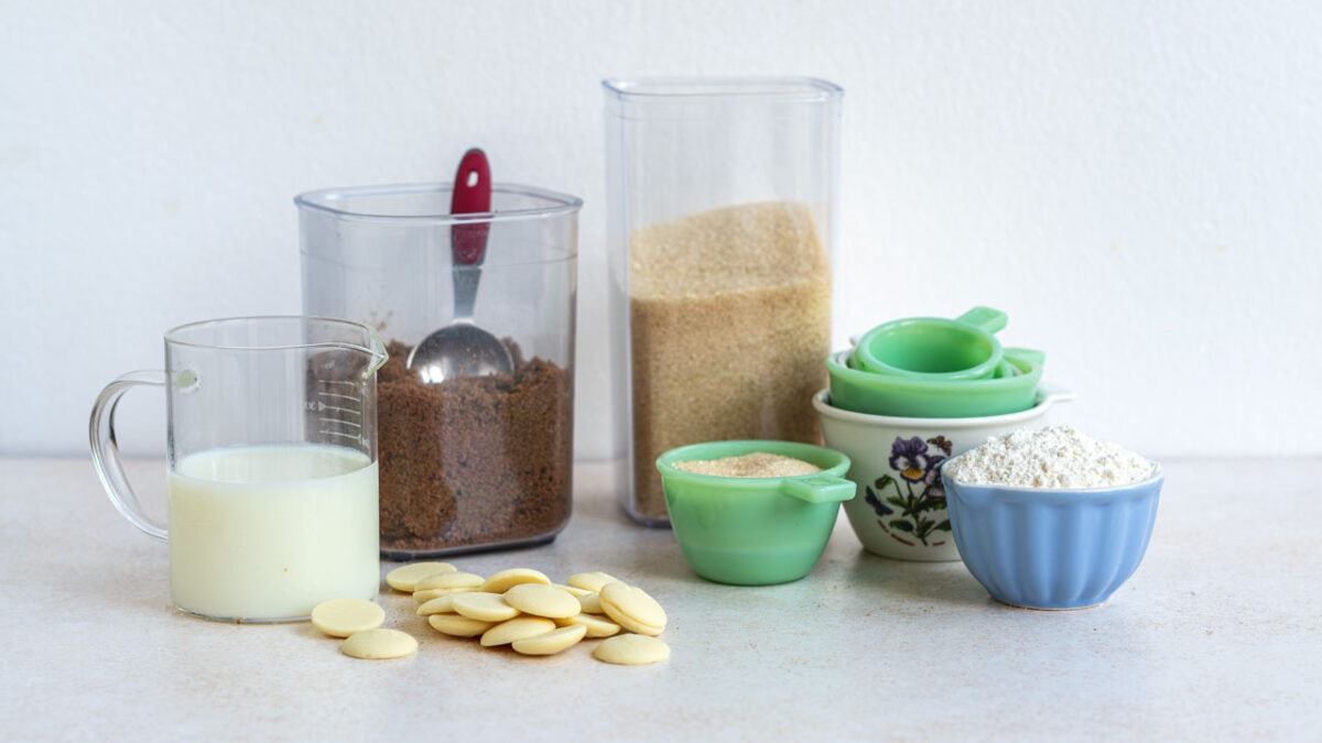 A variety of baking ingredients including milk, chocolate chips, instant coffee, sugar, flour and measuring cups arranged on a white countertop.