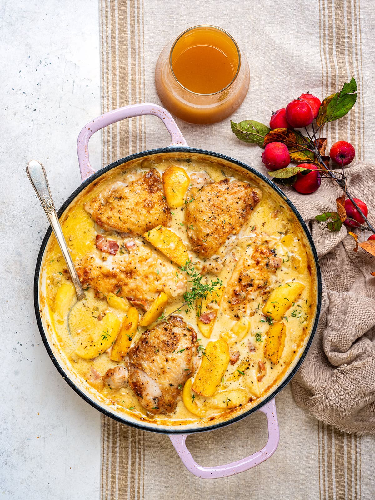 A pot of creamy chicken Normandy with apples and herbs sits on a striped cloth, next to a glass of cider, a beige napkin, and red apples with leaves.