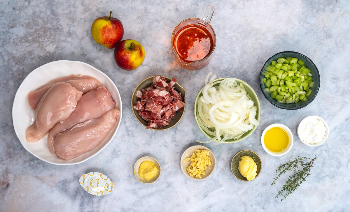 Ingredients for a recipe displayed on a countertop: raw chicken breasts, apples, diced bacon, sliced onions, chopped celery, apple juice, mustard, minced garlic, olive oil, butter, herbs, cornstarch, and a bouillon cube.