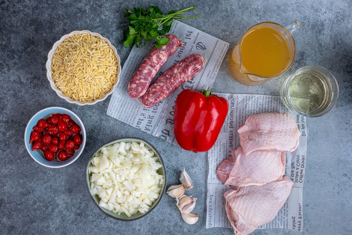 Ingredients for chicken scarpariello orzo laid out on a gray surface: orzo pasta, fresh parsley, two Italian sausages, bone-in chicken thighs, a red bell pepper, diced onion, a bowl of red drop peppers, garlic, a jug of chicken stock and a glass of white wine.