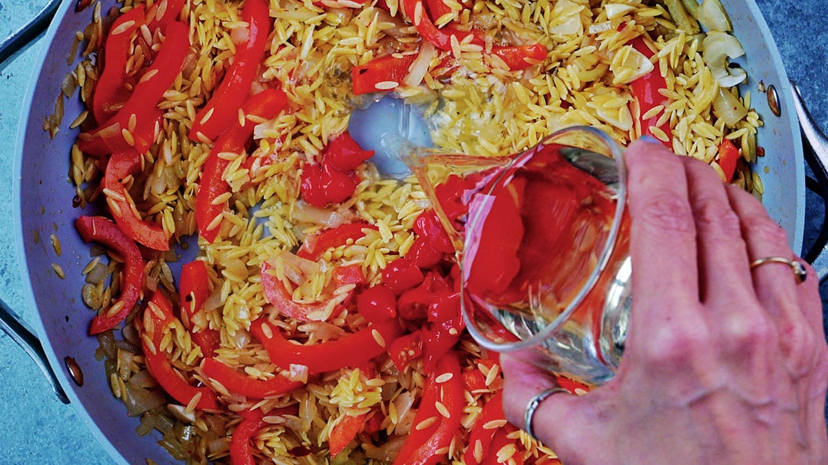 A hand pours white wine from a glass jug into a pan of toasted orzo with sauteed onion and red bell pepper strips.