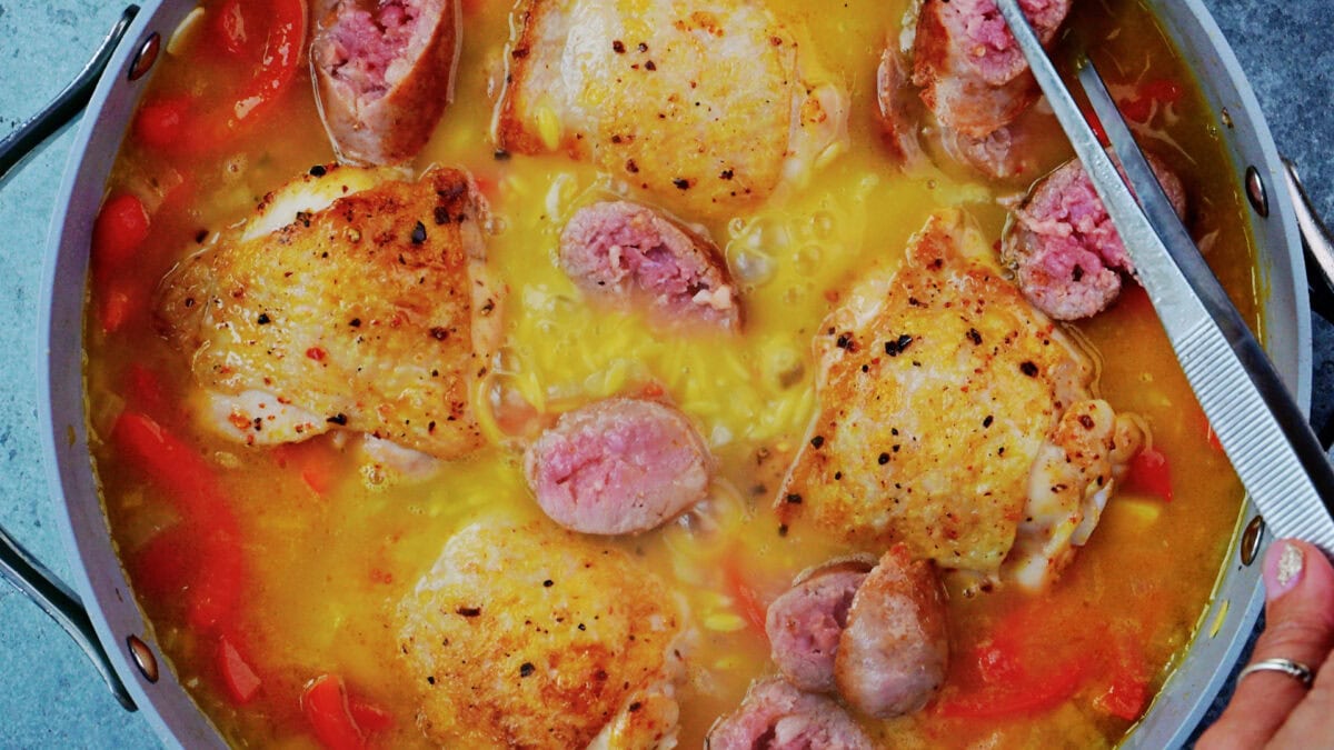 Seared chicken thighs set skin side up on orzo, sliced sausage and red bell peppers in a pan of chicken stock, ready to go into the oven.