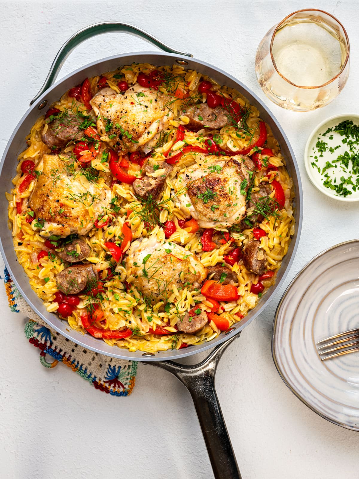 A skillet with roasted chicken thighs, sausage, orzo pasta, red bell peppers, and herbs sits next to a stack of plates with forks, a small bowl of chopped parsley, and a glass of white wine on a white surface.