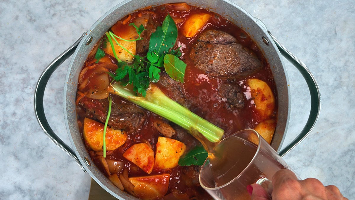 A hand pours liquid into a pot of beef stew with potatoes, celery, leafy herbs, and a rich tomato broth, all on a light countertop.