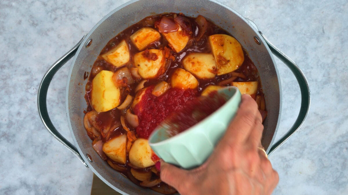 A hand pours tomato sauce from a light green bowl into a pot filled with potatoes, onions, and stew on a marble countertop.