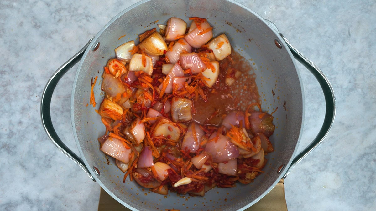 Diced onions and shredded carrots are sautéing in a large gray pot, with some liquid and seasonings visible at the bottom. The pot is placed on a light gray surface.