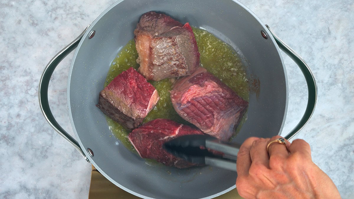 Four pieces of raw beef being seared in a gray pot with melted butter, as a hand uses tongs to hold one piece over the heat. The pot is on a white and gold surface.
