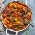 A white pot with Greek pot roast with tomatoes and potatoes, featuring tender beef stew, chunky potatoes, and a rich tomato sauce, garnished with chopped herbs. A spoon rests inside the pot, and a beige napkin is nearby on a gray surface.