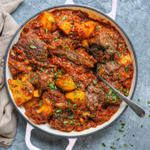 A white pot with Greek pot roast with tomatoes and potatoes, featuring tender beef stew, chunky potatoes, and a rich tomato sauce, garnished with chopped herbs. A spoon rests inside the pot, and a beige napkin is nearby on a gray surface.