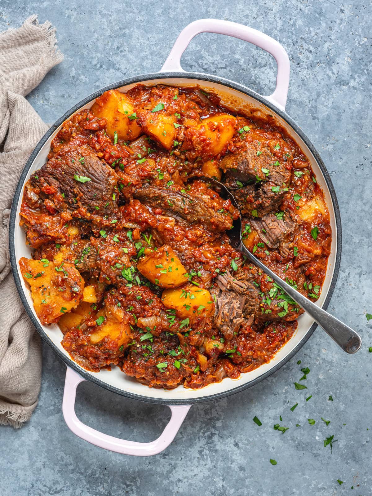 A white pot filled with beef stew, potatoes, and tomato sauce, garnished with chopped herbs. A spoon rests inside the pot on a gray background with a beige cloth nearby.