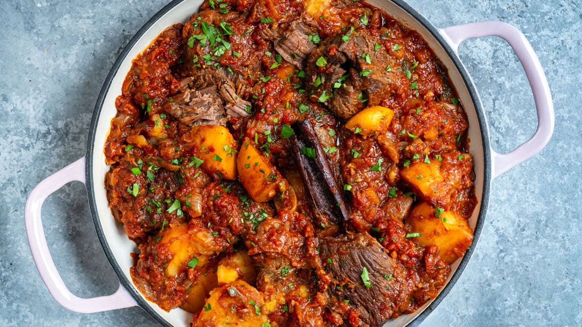 A pot of beef stew with chunks of beef, potatoes, tomato-based sauce, and fresh herbs, garnished with chopped parsley.