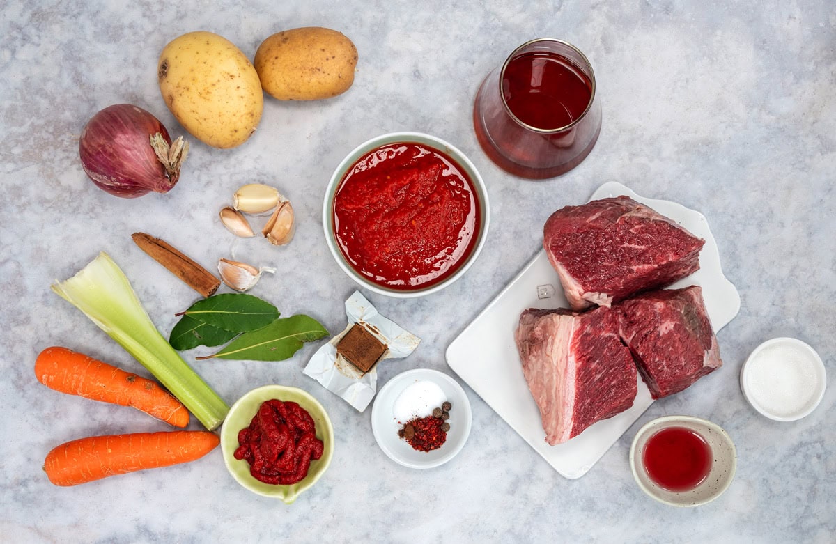 Raw ingredients for a stew, including potatoes, red onion, garlic, carrots, celery, beef chunks, tomato paste, tomato sauce, red wine, seasonings, and bay leaves arranged on a light surface.