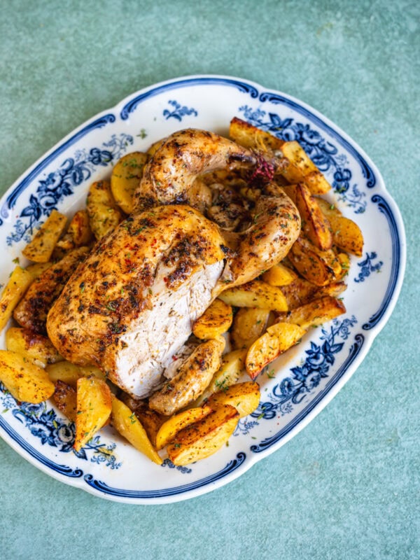 Greek roast chicken with crispy golden skin, partly carved, on a blue and white platter with lemon potatoes