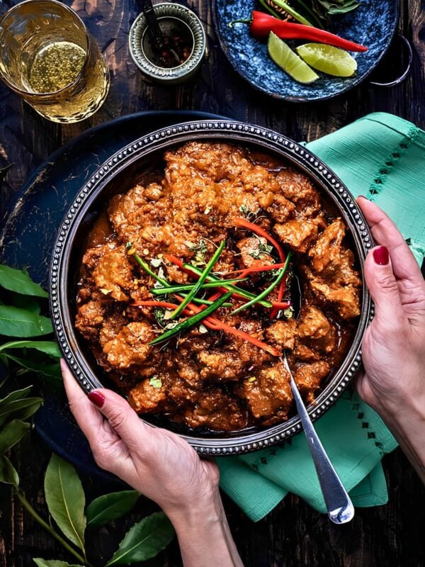 A hand holding a bowl of lamb bhuna garnished with sliced red chilies and herbs, with lime wedges, herbs and a drink on a dark wooden table.