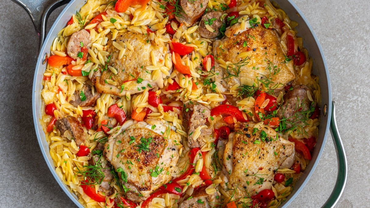 A skillet filled with golden-brown chicken thighs, sausage slices, orzo pasta, red bell peppers, red drop peppers, parsley and fennel fronds, all mixed together for a colorful, hearty meal.