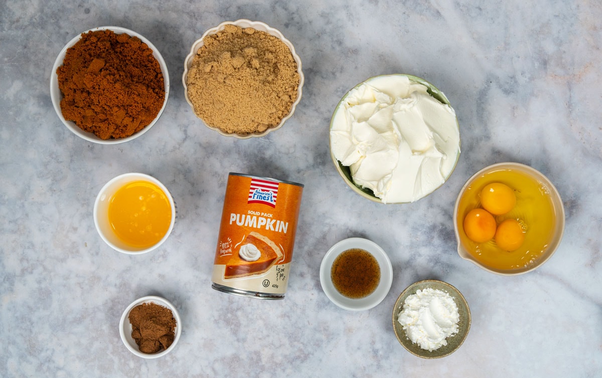 Ingredients for pumpkin pie cheesecake in bowls: crushed Biscoff cookies, melted butter, brown sugar, canned pumpkin purée, pumpkin pie spice, vanilla, mascarpone, eggs and cornstarch.