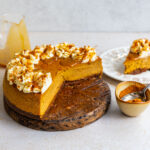 Pumpkin pie cheesecake with a Biscoff crust on a wooden board, a slice cut away to show the creamy filling, with a slice on a plate, a bowl of Biscoff crumbs and a jug of caramel sauce.