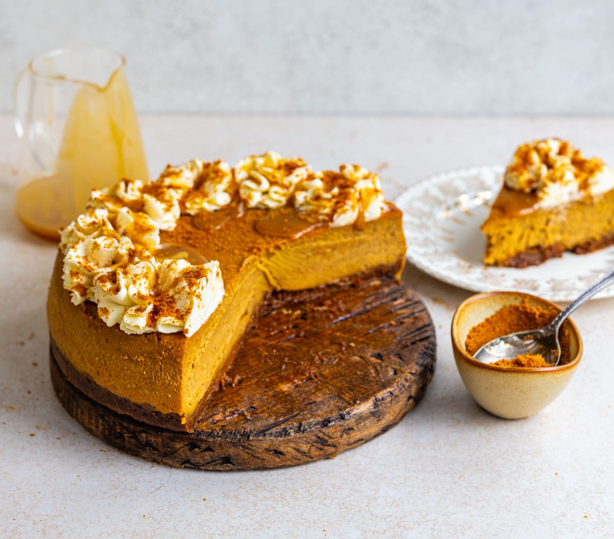 Pumpkin pie cheesecake with a Biscoff crust on a wooden board, a slice cut away to show the creamy filling, with a slice on a plate, a bowl of Biscoff crumbs and a jug of caramel sauce.