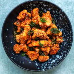 A black bowl filled with crispy, golden brown Taiwanese Fried Chicken pieces garnished with green herbs, set on a light blue textured surface.