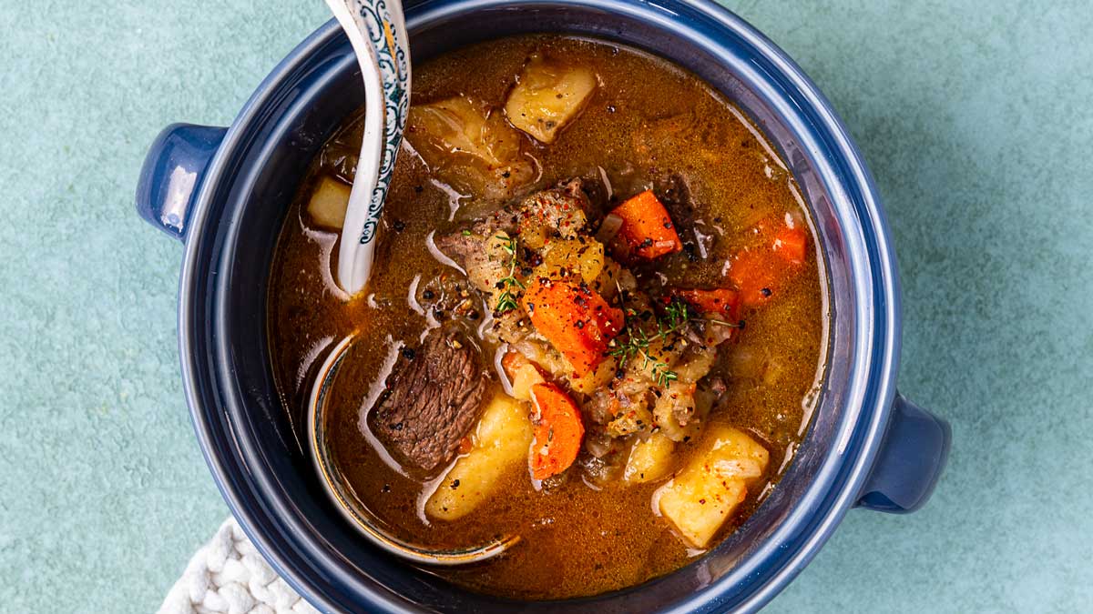 A blue pot of beef and vegetable soup with chunks of beef, carrots and potatoes and a patterned soup spoon.