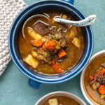 A blue bowl filled with beef stew containing chunks of meat, carrots, and potatoes, with a decorative spoon resting inside.