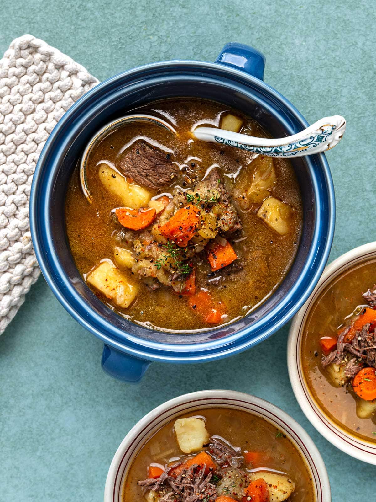 Overhead view of vegetable beef soup in a blue pot with a patterned spoon, with two smaller bowls of beef soup beside it.